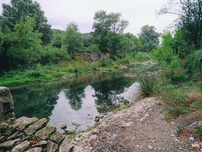 Photo d'un petit fleuve calme entouré d’arbres et de végétation, avec les pierres d'un ancien mur au premier plan et un reflet des arbres dans l’eau. Le ciel est très nuageux dans une faible lumière matinale.