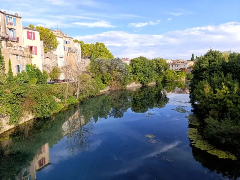 Vue d’un petit fleuve traversant un village. À gauche, des bâtiments aux façades colorées longent l’eau. À droite, des arbres et des berges herbeuses bordent le cours d'eau. Le ciel est dégagé avec quelques nuages.