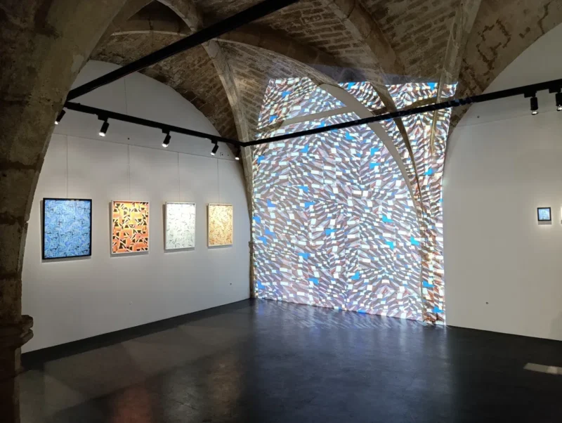 Photo d'une salle d’exposition avec voûtes en pierre, présentant cinq photos colorés accrochés au mur blanc et une grande projection lumineuse abstraite sur l’arc en fond.