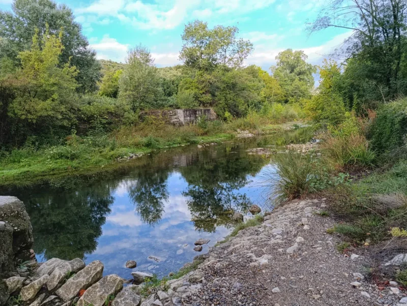 Photo d'un petit fleuve calme entouré d’arbres et de végétation, avec les pierres d'un ancien mur au premier plan et un reflet des arbres dans l’eau. Le ciel un peu nuageux dans une faible lumière matinale.