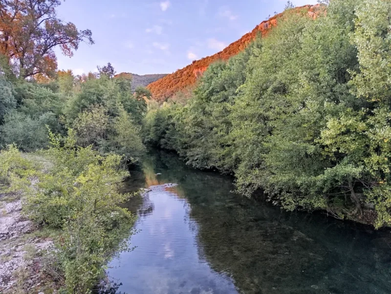 Photo d'un cours d'eau au petit matin entouré de végétation dense. De grands arbres aux feuilles luxuriantes bordent le cours d'eau sous un ciel bleu tacheté de petites tâches blanches. En arrière-plan, on aperçoit les collines boisées d'un petit massif karstique sous la lumière orangée du soleil levant.