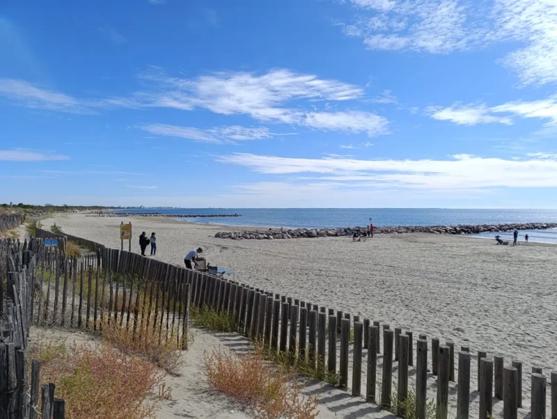 Photo d'une plage de sable bordée par la mer. Au premier plan, une clôture en bois traverse la dune, où poussent quelques herbes et buissons bas. Plusieurs personnes se trouvent sur la plage, certaines marchant, d’autres assises ou près de l’eau. Des digues en enrochement avancent dans la mer, découpant le rivage. Le ciel est bleu parsemé de nuages blancs.