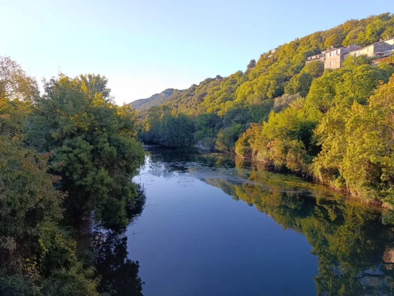Vue d’un petit fleuve bordé de collines boisées. À droite, des maisons en pierre sont visibles sur les hauteurs. Le ciel est dégagé mais la luminosité matinale encore faible.