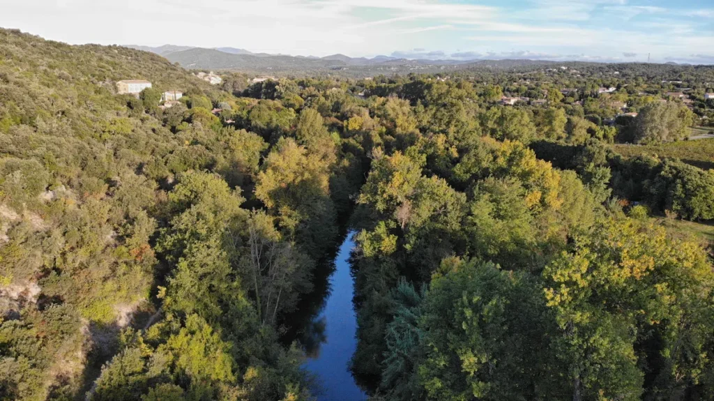 Vue aérienne d'un petit fleuve au milieu de la végétation. Plus loin, les maisons d'un village sous un ciel entre bleu et nuages d'altitude.