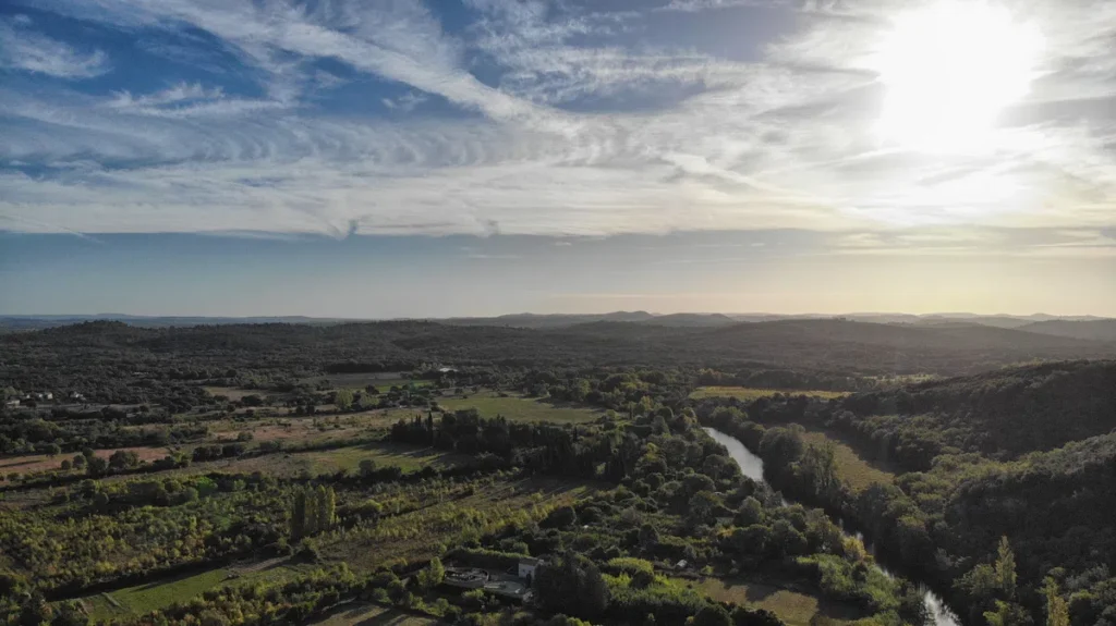 Vue aérienne d'un petit fleuve au milieu de la plaine. Au-dessus, le soleil du matin brille dans un ciel légèrement nuageux.