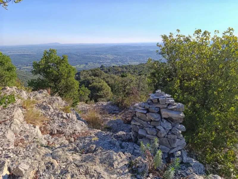 Photo d'un point de vue en hauteur sur un paysage gardois. Au premier plan, on distingue un tas de pierres empilées, entouré de rochers et de végétation basse. Autour, des arbustes et des arbres verts encadrent le tout. En arrière-plan, une vaste étendue de collines et de forêts s'étend jusqu'à l'horizon, sous un ciel dégagé et bleu.