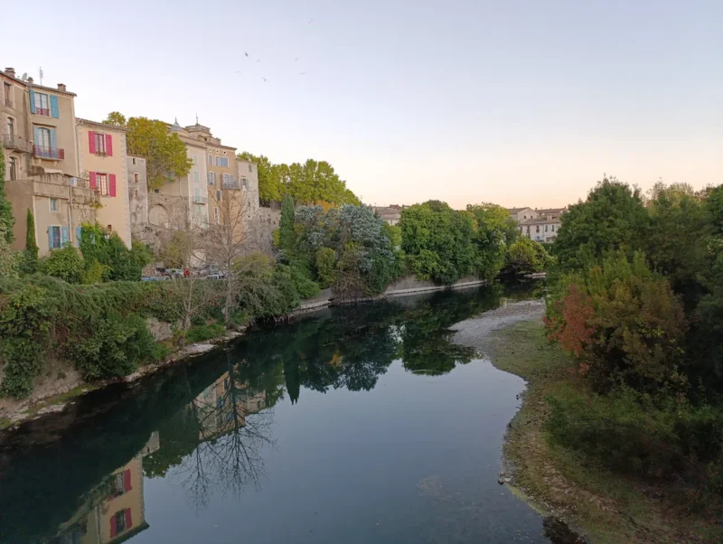Photo d'un petit fleuve bordé de végétation, avec des arbres verts et quelques feuillages aux teintes automnales sur la rive droite. Sur la rive gauche, on distingue une rangée de bâtiments anciens aux façades claires, dont certaines fenêtres ont des volets colorés (rouges et bleus). Leurs reflets apparaissent dans l’eau calme du Vidourle. À l’arrière-plan, on aperçoit d’autres habitations du petit village de Sauve surélevées. Le ciel du petit matin est clair, légèrement teinté de nuances pastel vers l’horizon.