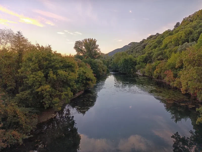 Vue d’un petit fleuve bordé de végétation. Le ciel est dégagé et se reflète dans l'eau mais la luminosité matinale encore faible.