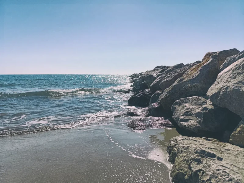Photo d'une plage de la Méditerranée. Eau calme avec petites vagues qui viennent se briser sur le rivage. À droite, une digue formée de gros rochers s’avance dans la mer. Le ciel est bleu sans l'ombre d'un nuage.