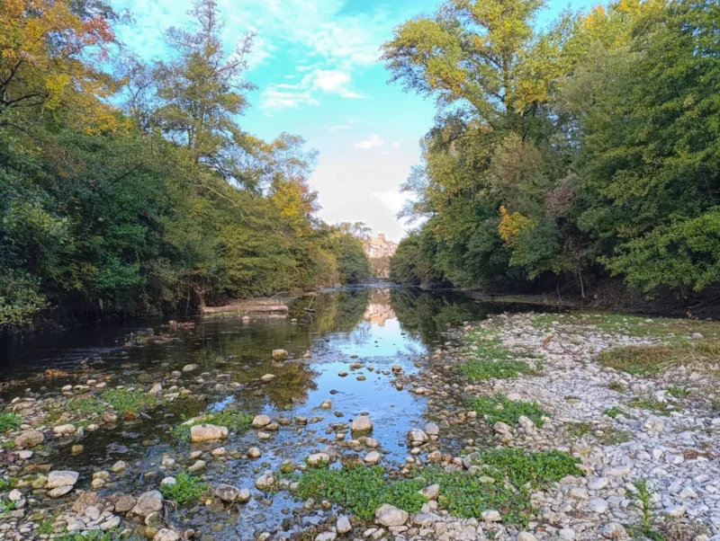 Photo d'un petit fleuve peu profond qui coule au centre de l’image, son lit parsemé de galets. De part et d’autre, des arbres denses aux feuillages verts bordent les rives, formant un couloir naturel. Au loin, on distingue quelques maisons. Le ciel est bleu mais tacheté de nuages.