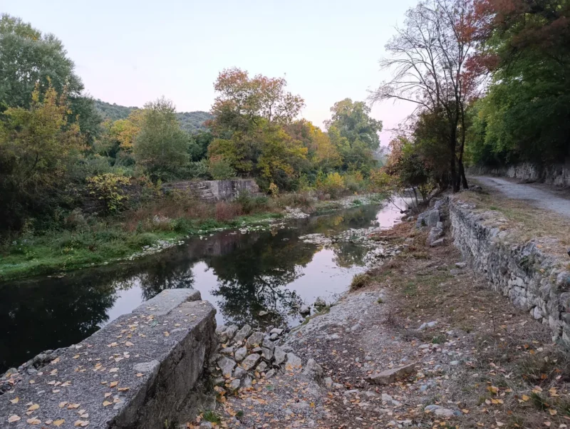 Photo d'un petit fleuve dont le niveau est bas, entouré d’arbres et de végétation, avec un ancien mur au premier plan et un reflet des arbres dans l’eau. Le ciel est dégagé dans une faible lumière matinale.