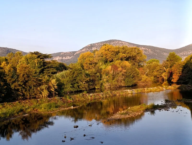 Au premier plan, les eaux d'un petit fleuve traversé dans sa largeur par une petite cascade artificielle. Derrière, une rangée d'arbres aux couleurs automnales. Derrière, un petit massif sous un ciel bleu.