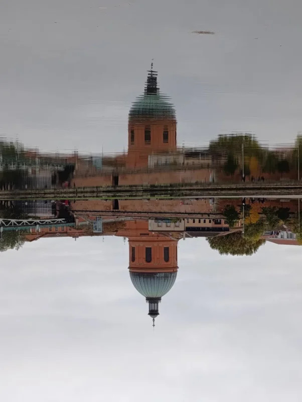 Photo du reflet de la chapelle Saint-Joseph de la Grave, un bâtiment à dôme vert dans l’eau de la Garonne, créant une symétrie verticale nette mais légèrement déformée par les ondulations de la surface. Le ciel est gris uniforme et les rives sont bordées de constructions en briques orangées et de quelques arbres aux feuillages d’automne. L'image a été retournée de sorte que le reflet est en haut de l'image.