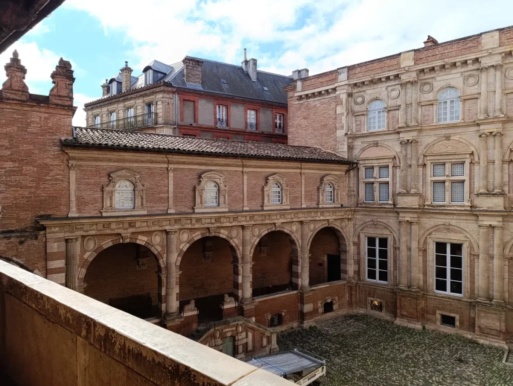 Cour intérieure d'un bâtiment historique en brique avec des galeries à arcs et des fenêtres classiques, entourée d'architectures anciennes sous un ciel partiellement nuageux.