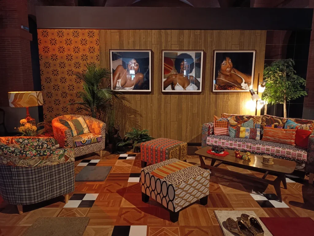 Photo d'un salon reconstitué au style rétro : canapé, fauteuils et poufs aux motifs colorés, table basse, lampes, plantes vertes et trois portraits de femmes accrochés au mur en bois.