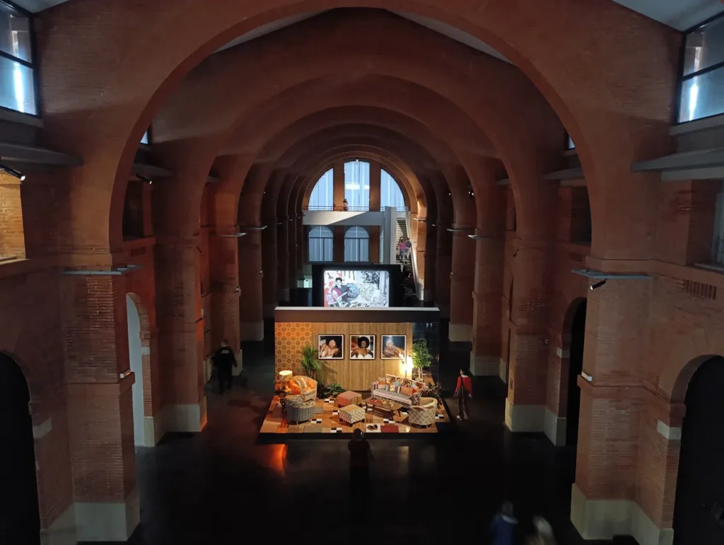 Vue intérieure d'une salle d'exposition des Abatoirs à Toulouse, en brique avec des arches, présentant une installation artistique centrale composée de meubles, d'un écran et de portraits encadrés.
