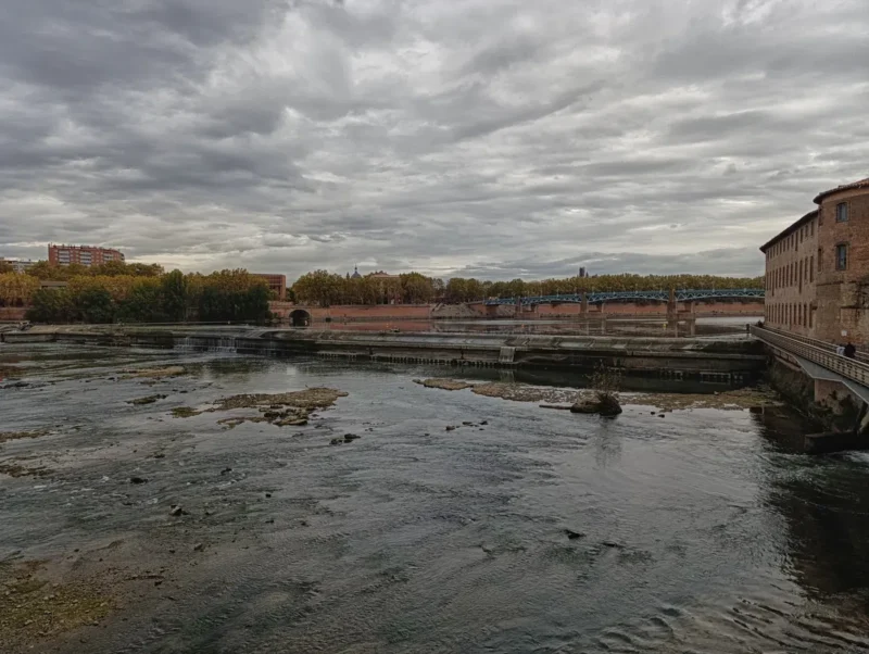 La Garonne à Toulouse sous une épaisse couche nuageuse. On distingue des bâtiments de briques rouges sur la droite et un pont qui enjambe le fleuve.