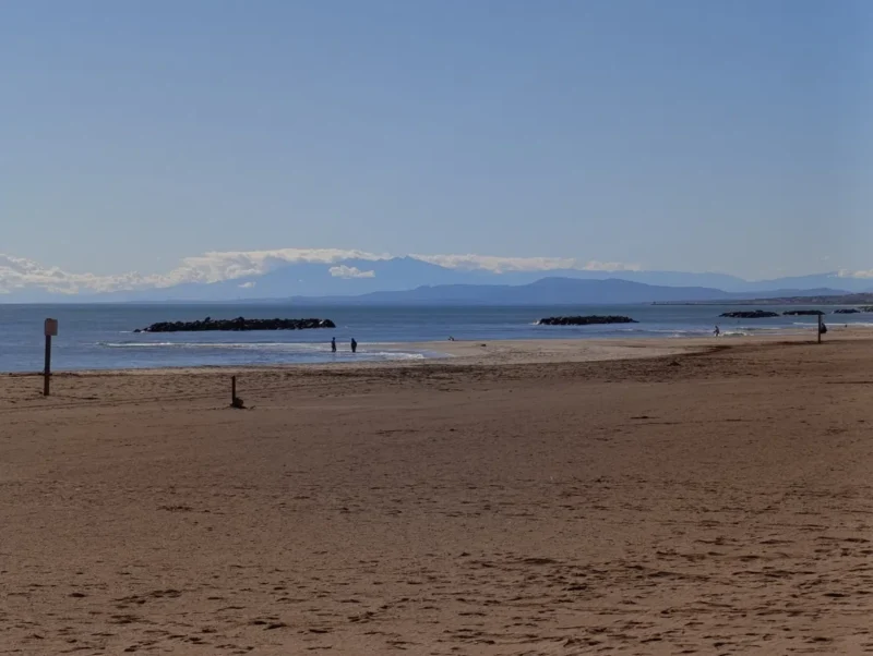 Photo d'une plage quasi déserte. Vient ensuite la mer avec des digues de roches parallèles au littoral et, au loin, des montagnes avec des nuages. Au-dessus, le ciel est bleu.