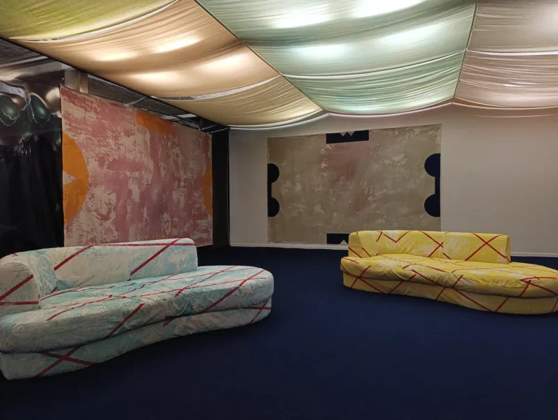 Photo d'un espace d'exposition avec des canapés colorés et des toiles abstraites accrochées aux murs, sous un plafond tendu aux tons pastel.