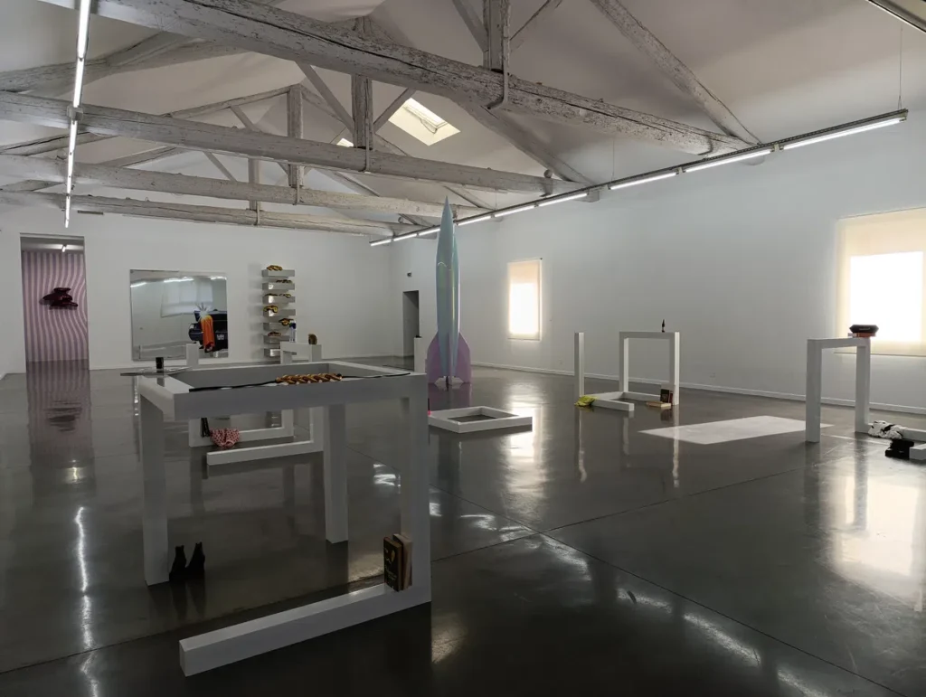 Photo d'une salle d'exposition spacieuse et lumineuse avec des structures blanches, une fusée bleue suspendue et des objets artistiques disposés sur des tables basses.