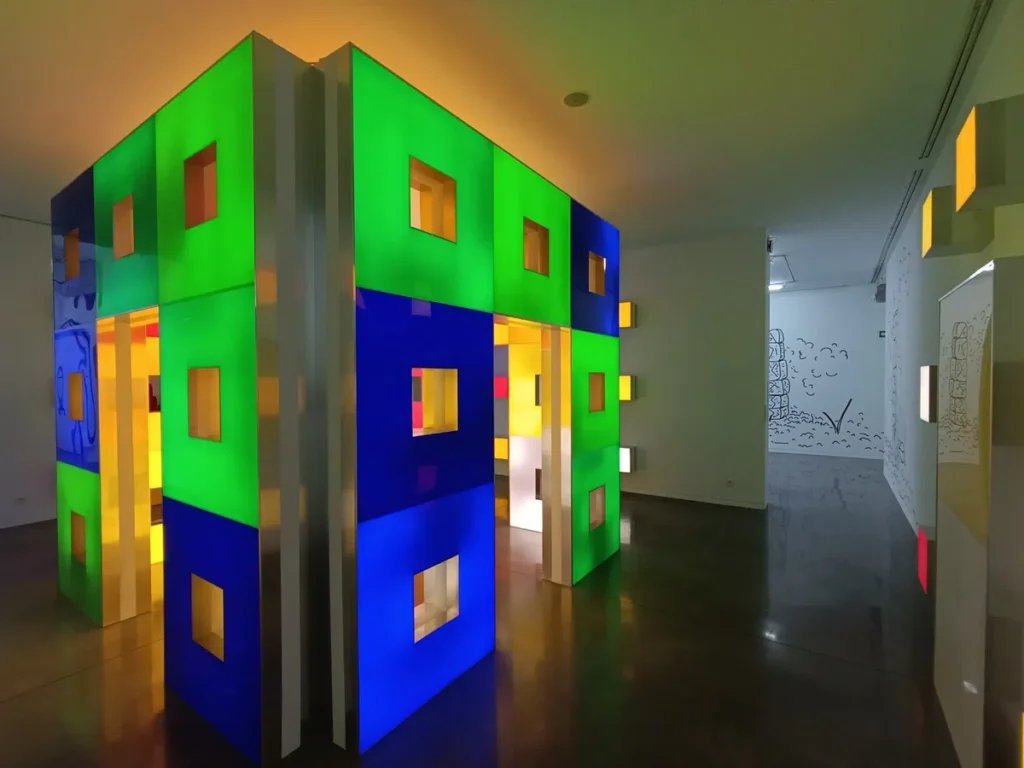 Photo d'une installation artistique lumineuse et colorée en forme de labyrinthe, avec des murs aux tons verts, bleus et jaunes.