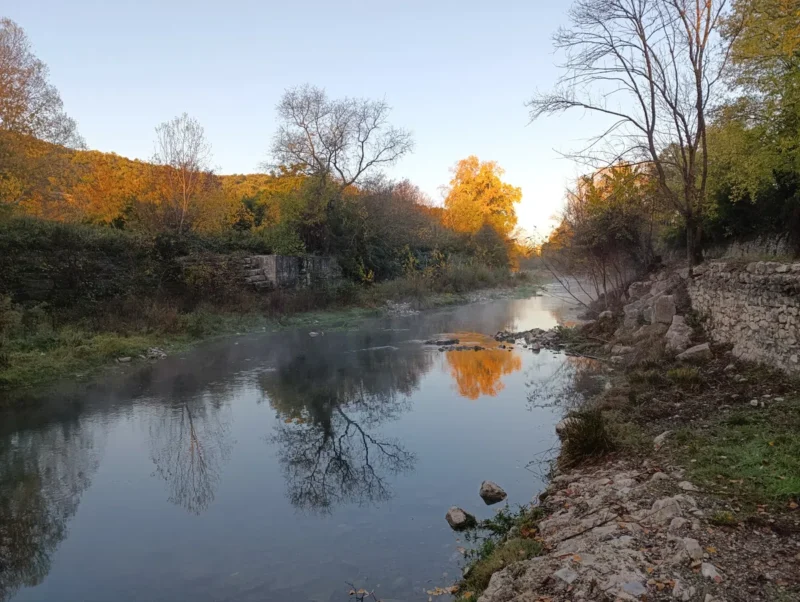 Photo d'un petit fleuve calme, entouré d’arbres et de végétation, avec un vieux mur de pierre sur la droite et un reflet des arbres dans l’eau. Le ciel est dégagé et une légère brume flotte au-dessus de l'eau dans une faible lumière matinale.