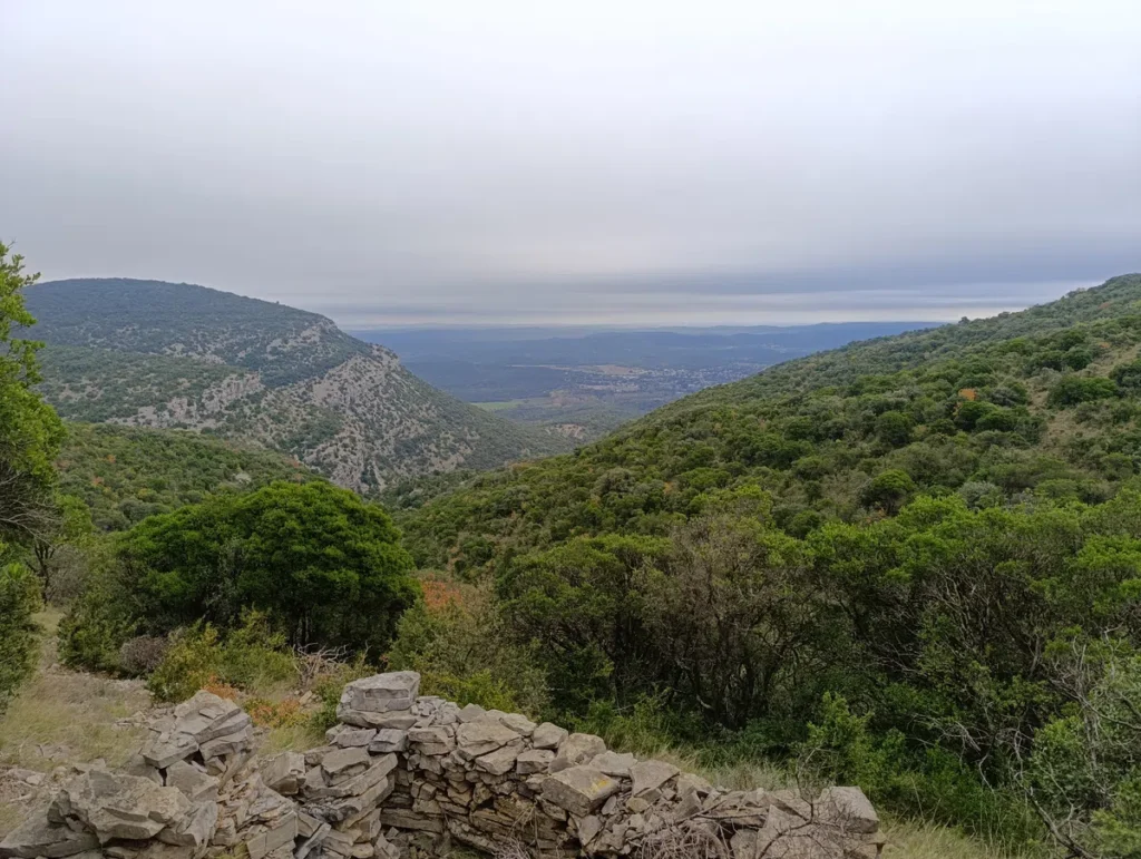 Photo prise du fond d'une reculée karstique. Derrière une petite ruine, au premier plan, un paysage naturel de collines verdoyantes avec une vue dégagée sur une plaine sous un ciel nuageux.
