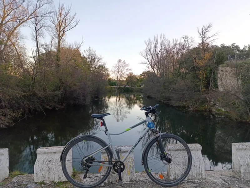 Vélo posé contre une rambarde en béton sur un petit passage à gué, au-dessus d’un petit fleuve calme bordé d’arbres dénudés. Ciel dégagé dans une faible lumière matinale.