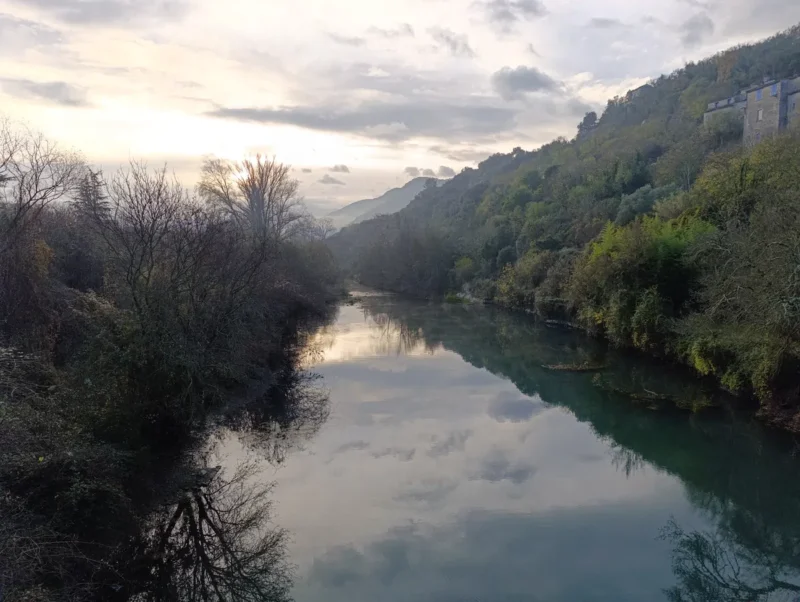 Un petit fleuve entouré de végétation, avec un massif karstique boisé en arrière-plan et un ciel partiellement nuageux reflétant à la surface de l'eau.