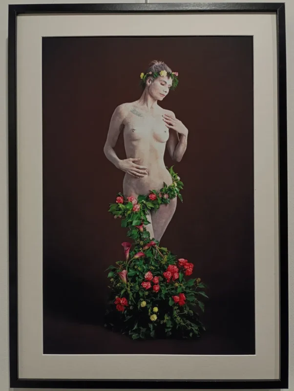 Photographie encadrée montrant une femme nue posant debout, le corps partiellement couvert de feuillage et de fleurs colorées, avec une couronne florale sur la tête, sur fond uni sombre.