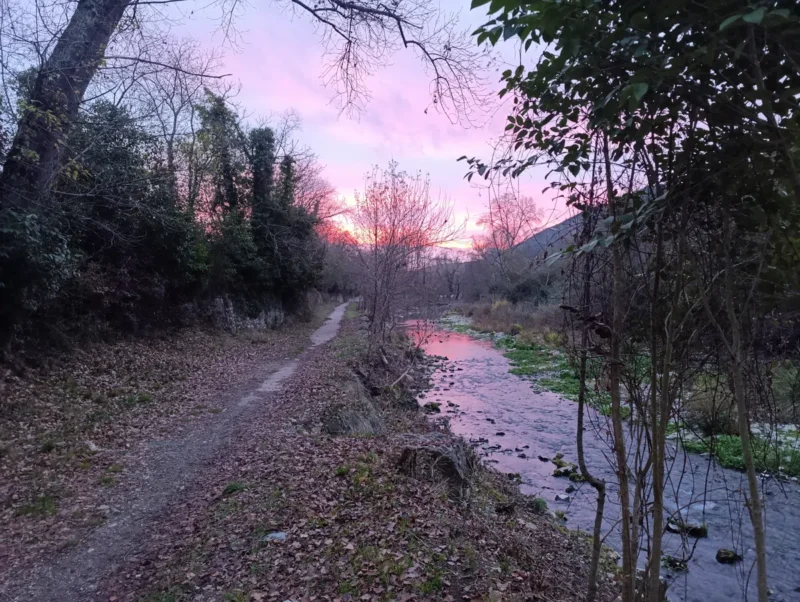 Un sentier le long d'un petit fleuve dans lequel se reflètent les couleurs du ciel illuminé par la lumière du soleil levant. Ciel partiellement nuageux.