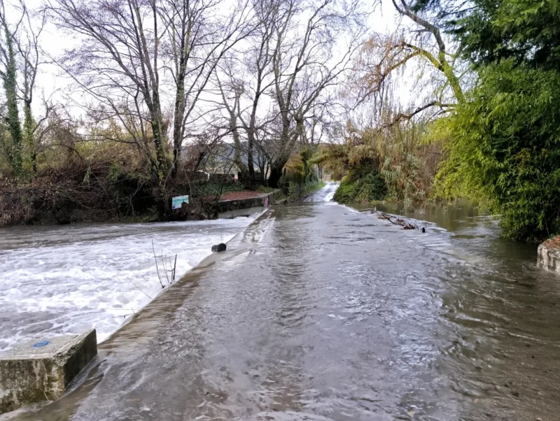 Au premier plan, un passage à gué sur lequel le cours d'eau tumultueuse est passé sur moins d'une dizaine de centimètres. La route n'est plus praticable. Le petit fleuve est bordé d'arbres.