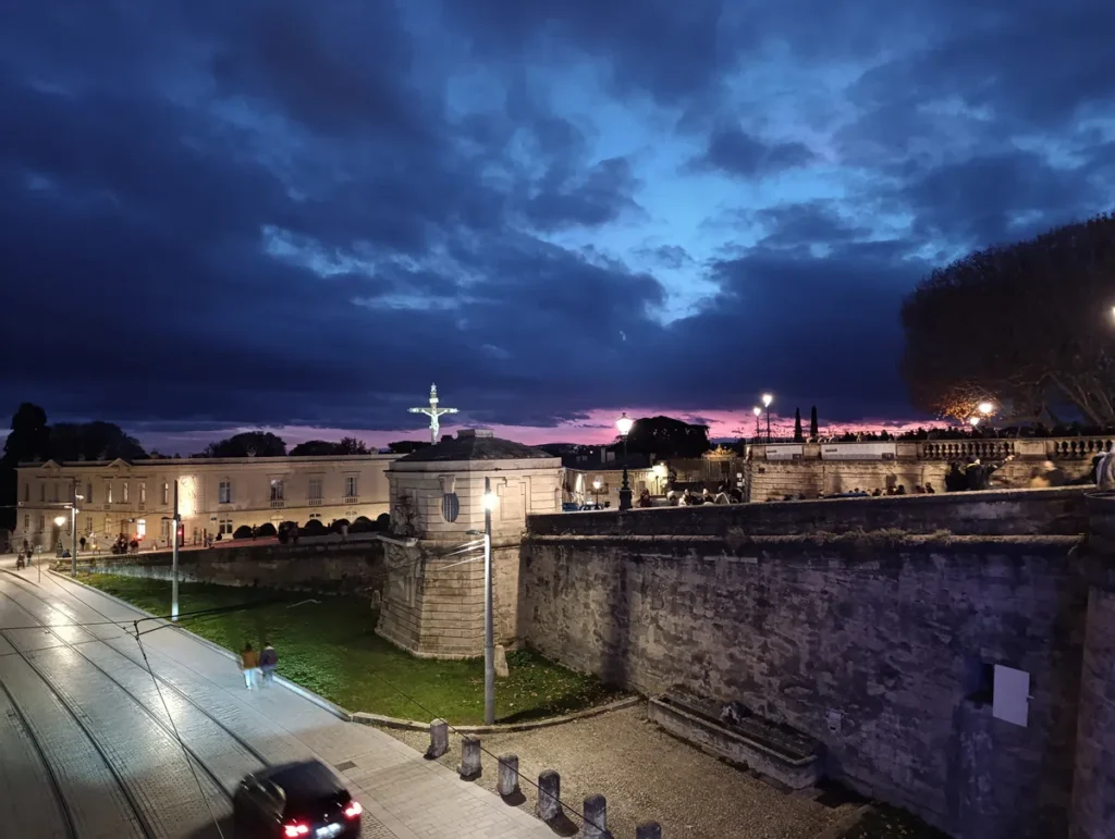 Vue de ce qui pourrait sembler une fortification de pierres au crépuscule, le Pérou, à Montpellier, avec une rue éclairée au premier plan, une voiture un peu floue, des piétons et une grande croix éclairée posée derrière une petite tour, sous un ciel nuageux teinté de bleu et de violet.