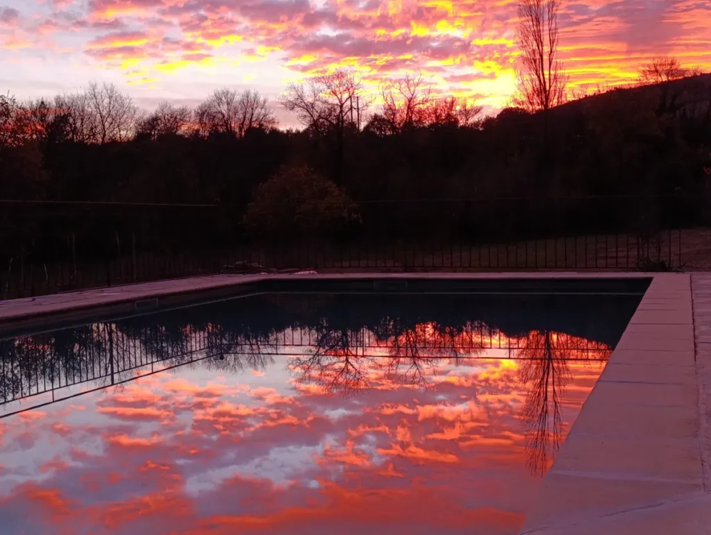 Un ciel de soleil couchant, semblable à un incendie, se reflète dans l'eau d'une piscine, au premier plan. Au loin, sous le ciel, une rangée d'arbres dans l'obscurité.