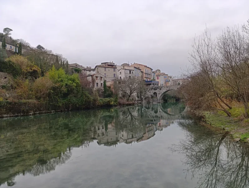 Un village sur la rive opposée d'une petite étendue d'eau, un petit fleuve dont un petit barrage freine le cours. Un pont enjambe le fleuve. Le ciel est gris au-dessus.