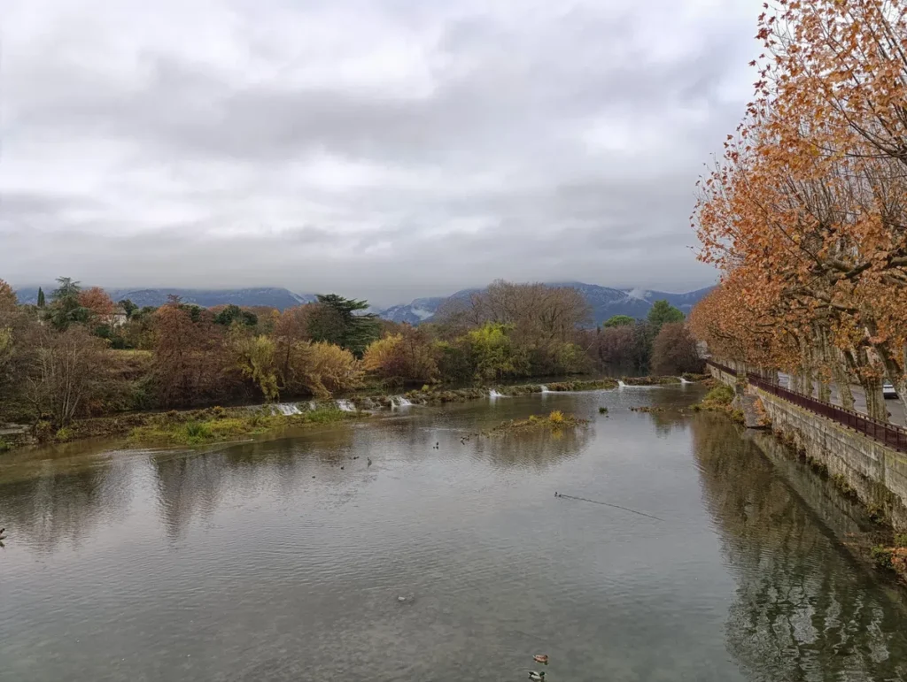 Un cours d'eau avec une rangée de platanes sur la droite de l'image. Une petite cascade artificielle traverse le fleuve dans sa largeur. Au fond, un petit massif sous un ciel très nuageux.