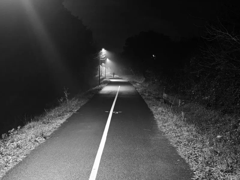 Une voie verte de nuit dans la lumière des lampadaires et dans une légère brume. Au loin, une personne marche.