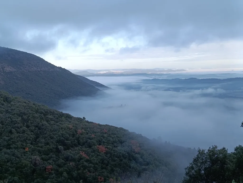 Sous un ciel voilé de nuages, une large nappe de brume vue des hauteurs d'un massif dont on distingue les pentes sur la gauche de l'image.