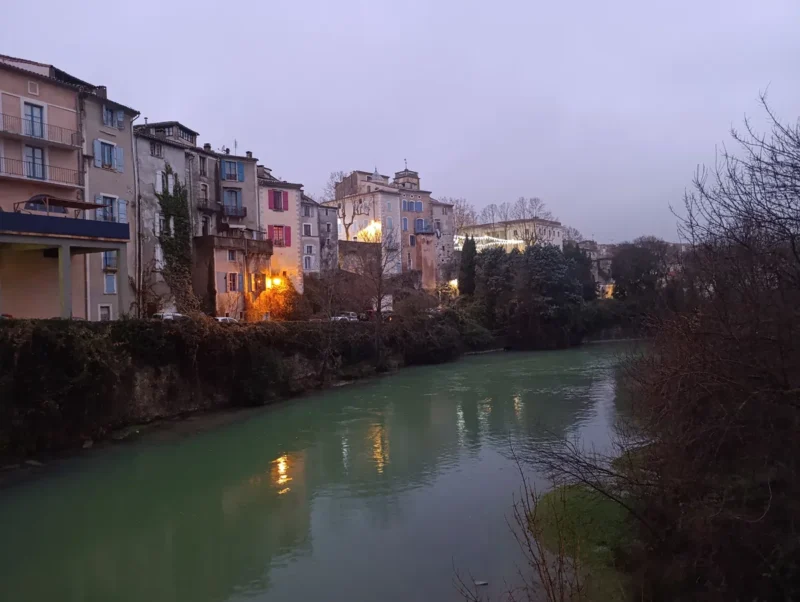 Un village au bord d'un petit fleuve à l'eau boueuse. La lumière matinale est faible et on distingue éclairage et décorations lumineuses de Noël. Le ciel est très nuageux.
