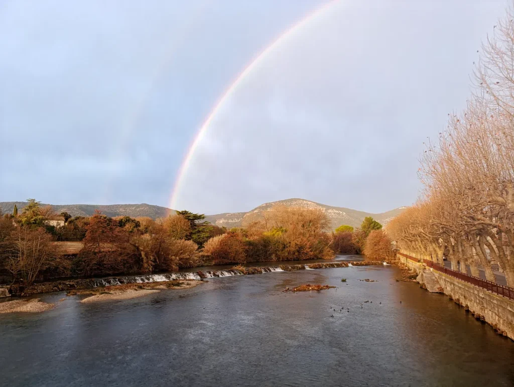 Photo d'un arc-en-ciel au-dessus d’un petit fleuve avec une petite cascade artificielle, des platanes alignés sur les berges, côté droit de l'image, et petit massif en arrière-plan.