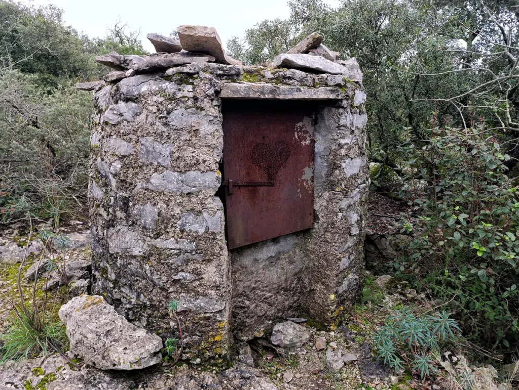 Un vieux puits en pierre avec une porte métallique rouillée, entourée de végétation méditerranéenne.
