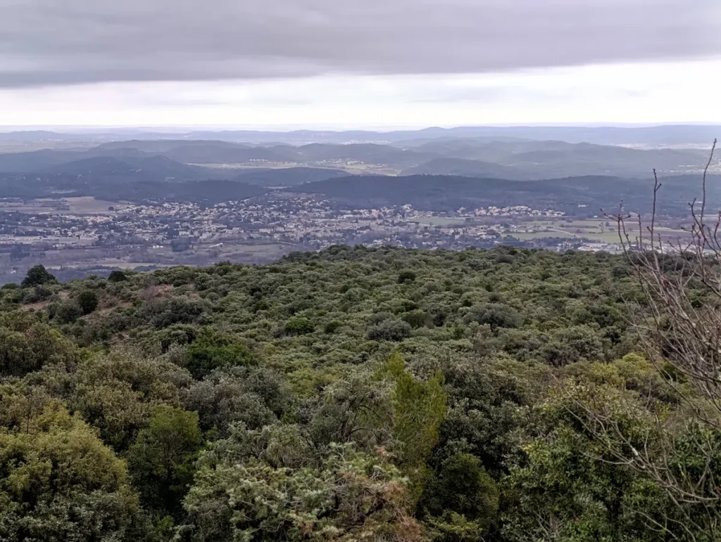 Sous un ciel couvert, photo prise depuis les hauteurs d'un massif. On distingue à mi-plan, dans la vallée, les maisons d'une petite ville. Devant, la végétation couvre le haut du massif. En arrière-plan, le paysage s'étend sur un relief de collines.