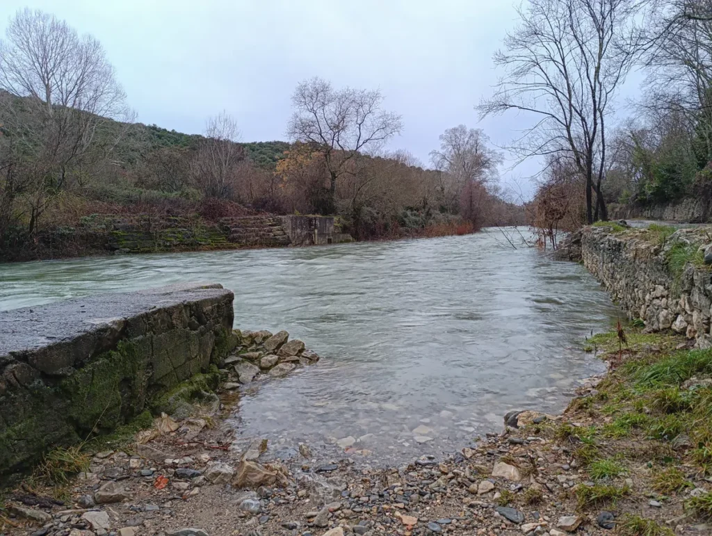 Photo d'un petit fleuve à l'eau tumultueuse bordé d’arbres et de végétation, avec un ancien mur au premier plan. Le ciel est très nuageux dans la lumière matinale.
