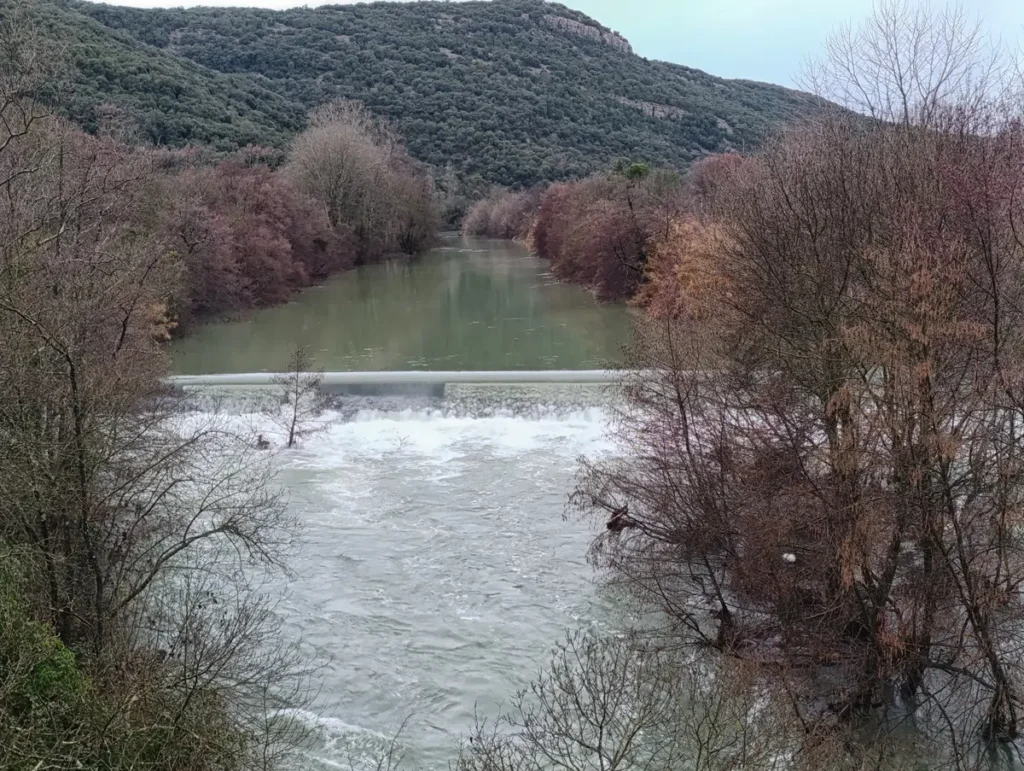 Au pied d'un massif, un fleuve à l'eau tumultueuse, au niveau d'une petite cascade qui permettait de canaliser l'eau d'un moulin. Le fleuve est bordé de végétation en mode hivernal. Ciel couvert, à peine visible.