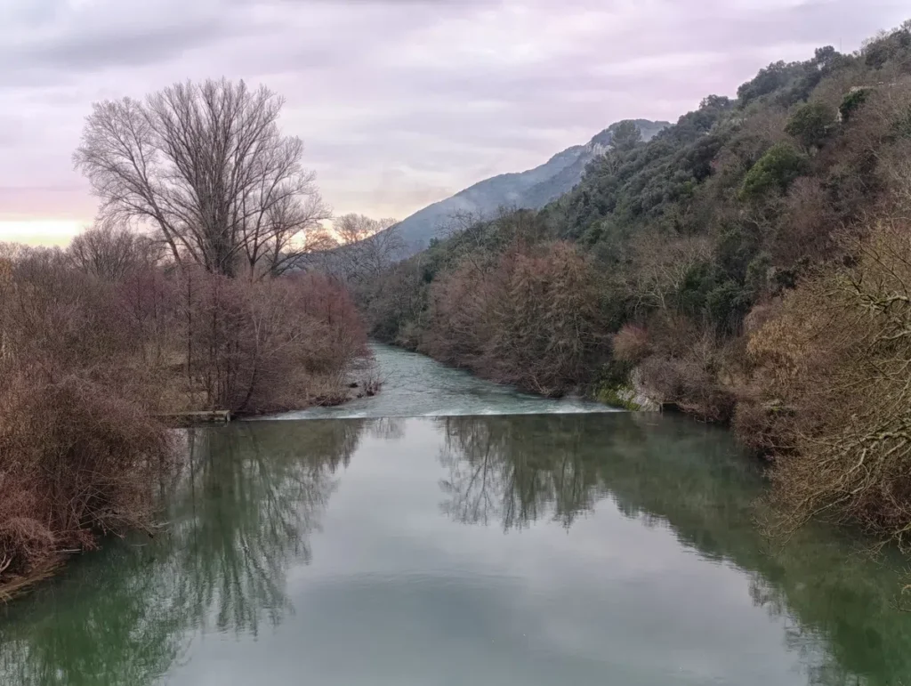 Vue d’un petit fleuve bordé de collines boisées. Une petite cascade artificielle entrave le cours du fleuve. Le ciel est couvert et la végétation hivernale.