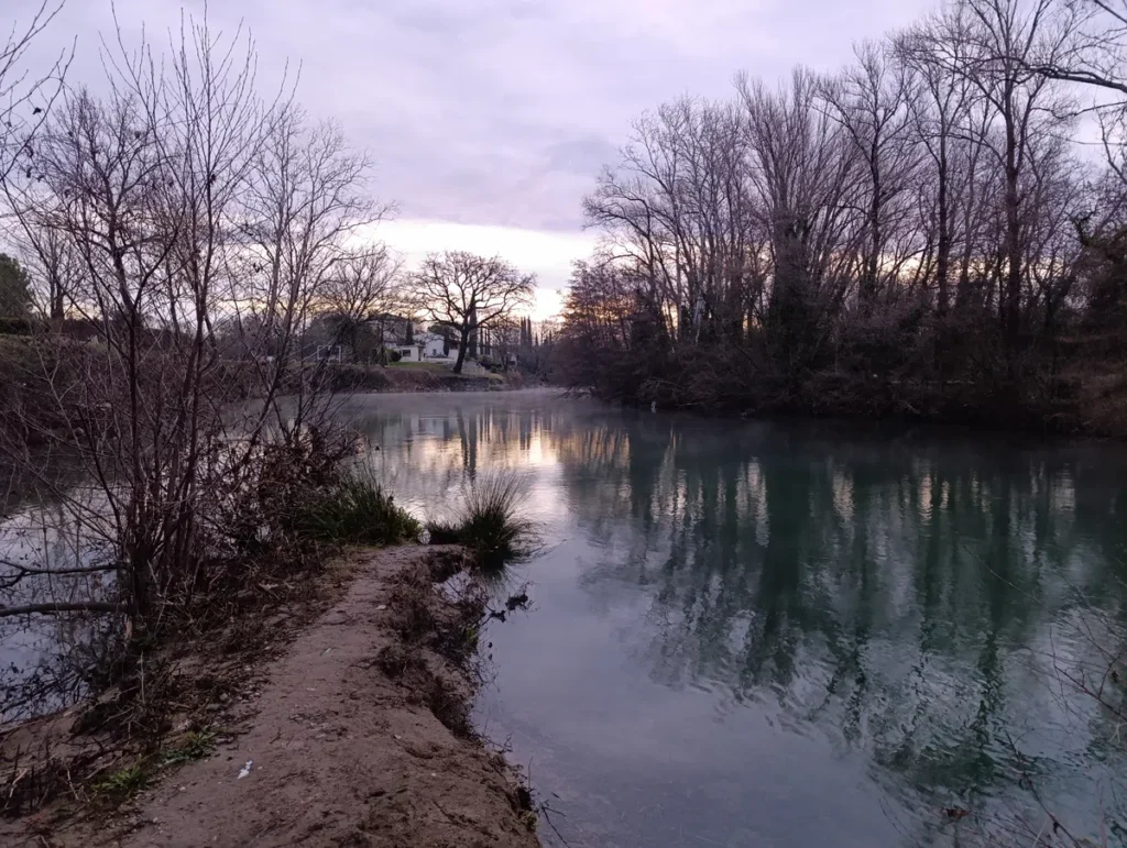 Photo d'un paysage avec un petit fleuve à l'eau calme bordée d'arbres principalement dépourvus de feuilles. Un sentier étroit avance vers l'eau au premier plan. La rivière reflète les arbres et le ciel. À l'arrière-plan, une maison claire avec un toit sombre est visible derrière une petite butte herbeuse, partiellement cachée par des arbres. Le soleil est bas et partiellement masqué par les branches, projetant une lumière diffuse.