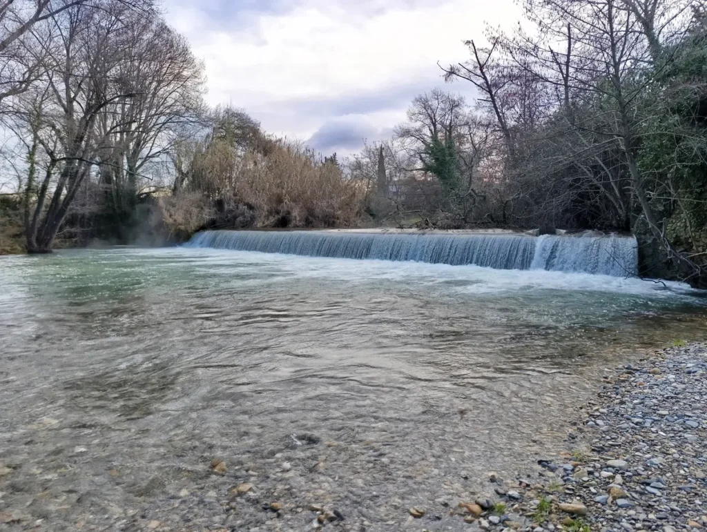 Une petite cascade artificielle derrière une plage de galets. Au fond, des arbres sous un ciel nuageux.