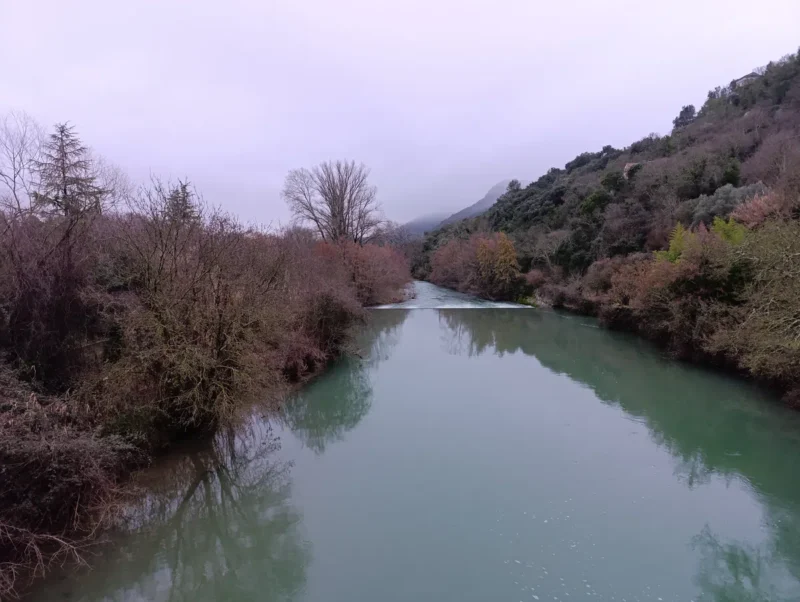 Photo prise d'un pont surplombant les eaux verdâtres d'un petit fleuve aux rives arborées à la végétation hivernale, sous un ciel bas qui masque les hauteurs d'un massif dont on perçoit un bout sur la droite de l'image.