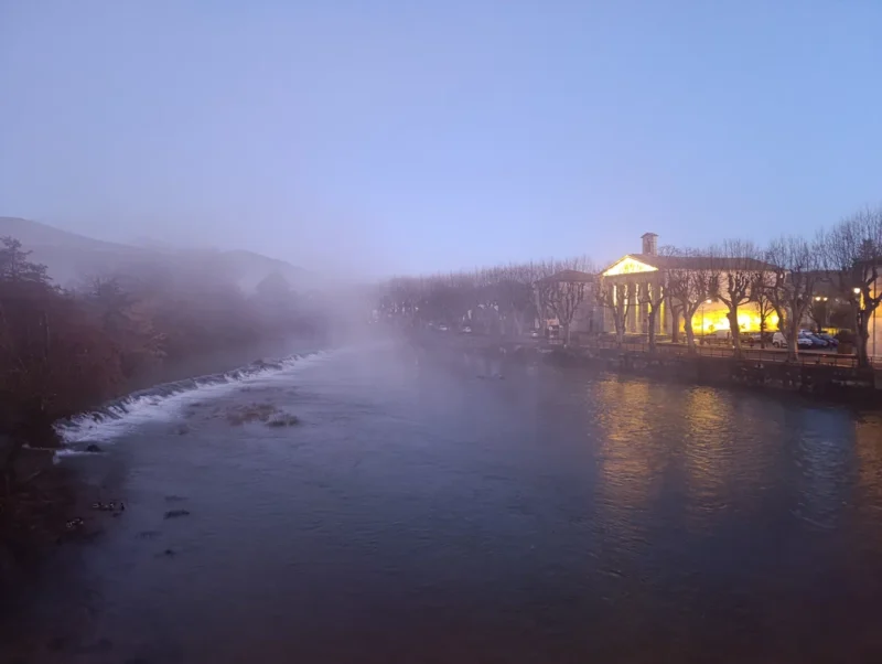 Photo prise d'un pont qui surplombe un petit fleuve, dans une faible lumière matinale. Sur la droite, un temple éclairé par des lumières artificielles. Une cascade coupe le cours d'une rive à l'autre. Une brume s'étend au-dessus de l'eau, bien qu'au-dessus le ciel soit dégagé.