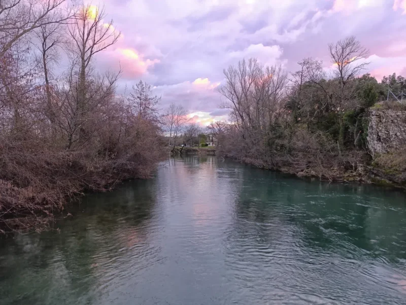 Photographie d’un cours d’eau bordé de végétation. Sur les deux rives, on observe des arbres partiellement dénudés. Le ciel est nuageux dans la lumière colorée du matin.